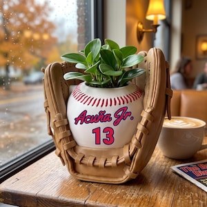 Peut inclure: Un gant de baseball en cuir beige tenant un pot en céramique blanc en forme de balle de baseball avec des coutures rouges. Le pot porte l'inscription Acuña Jr. et le numéro 13 en bleu et rouge, contenant une petite plante verte aux feuilles rondes.