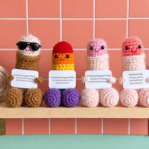 May include: Four crocheted phallic figures in various colors, each holding a white card with text. The first is brown with sunglasses, the second is a rainbow gradient, the third is light pink, and the fourth is a darker pink with a smiling face.
