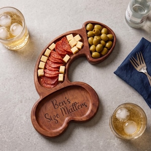May include: A phallus-shaped wooden serving board with three distinct compartments. The board features the engraved text "Ben's Size Matters" and is filled with sliced salami, cubed cheese, and green olives, accompanied by two glasses of iced drinks.