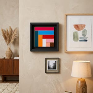 Könnte beinhalten: Ein quadratisches Wandkunstwerk in einem schwarzen Schattenfugenrahmen mit einer geometrischen Komposition aus rechteckigen Blöcken in Rot, Blau, Orange, Weiß, Rosa, Braun und Türkis, präsentiert neben einem gerahmten Druck und einem kleinen Foto.