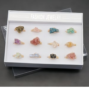 Puede incluir: Una caja de presentación blanca con la etiqueta FASHION JEWELRY que contiene doce anillos. Cada anillo presenta una piedra preciosa en bruto de forma irregular envuelta en un fino alambre dorado. Las piedras varían en color, incluyendo tonos de verde, rosa, azul, morado, rojo, transparente, amarillo y negro, dispuestas en una cuadrícula de tres por cuatro.