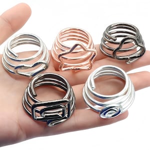 Puede incluir: Cinco anillos de metal con un diseño en espiral tipo resorte. Cada anillo presenta una forma decorativa única en el extremo de la espiral, incluyendo un corazón, una estrella, una nube, un cuadrado y un círculo. Acabados en plata, oro rosa y negro.