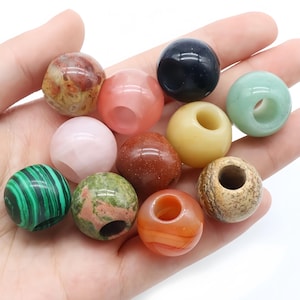 Könnte beinhalten: Eine Sammlung von elf kugelförmigen Perlen aus verschiedenen Natursteinen und Materialien, die eine Vielfalt an Farben aufweisen, darunter grüner Malachit, gesprenkeltes Braun, durchscheinendes Rosa, sattes Schwarz, blasses Gelb und orangefarbene Bänder.