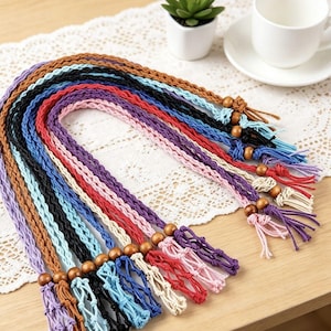 May include: A collection of braided cord lanyards in various colors including brown, light blue, black, red, pink, purple, and cream. Each lanyard features a wooden bead accent near the end and terminates in a knotted tassel fringe design.
