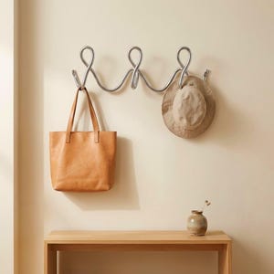 Könnte beinhalten: Eine silberfarbene Wandgarderobe aus Metall mit drei durchgehenden Schlaufen, an deren linkem Haken eine beigefarbene Ledertasche und an deren rechtem Haken ein beigefarbener Stoffhut hängt, montiert an einer schlichten Wand.