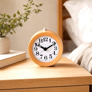 Peut inclure: Un réveil analogique rond avec un cadre en bois clair et un cadran blanc. L'horloge présente des chiffres arabes noirs de 1 à 12, des aiguilles des heures et des minutes noires, ainsi qu'un petit bouton argenté sur le dessus du boîtier en bois.