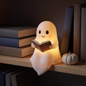 May include: A glowing white ghost-shaped lamp with a smooth surface, black eyes, and a small smile, holding an open brown book. The lamp emits a warm light and is positioned on a wooden shelf next to a small white pumpkin and several books.