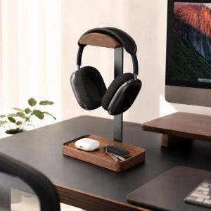 May include: A dark wood headphone stand with a vertical support and a rectangular base tray. Black over-ear headphones rest on the top curved wooden support. A white wireless earbud case and a set of metal keys are placed on the wooden base tray.