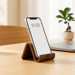 Può includere: Un supporto per smartphone in legno marrone scuro con una forma curva a onda. Il supporto sostiene uno smartphone bianco in posizione verticale. Lo schermo del telefono mostra l'ora 10:09 in cifre digitali nere su uno sfondo bianco.