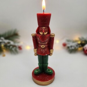 May include: A red and green nutcracker-shaped candle with gold accents on the hat, shoulders, belt, and base. The candle is lit, featuring a bright flame at the top of the hat. The figure has a molded mustache and uniform details in wax.