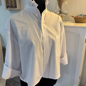 May include: A crisp white button-down shirt featuring a high ruffled collar, long sleeves with folded cuffs, and a single chest pocket. The garment is displayed on a black mannequin, showcasing a front button placket with five visible white buttons.
