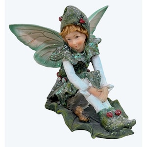 Peut inclure: Une figurine de fée aux cheveux châtain clair, portant une tunique verte à capuche ornée de baies rouges et de paillettes. Elle possède de grandes ailes vertes translucides, des manches blanches et des bottines vertes, assise sur un socle en forme de feuille.
