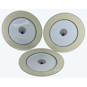 May include: Two round ceramic plates and one oval ceramic platter, each featuring a white center with a dark blue seashell illustration, a dark blue inner rim, and a wide cream-colored border. The set displays a consistent nautical design.