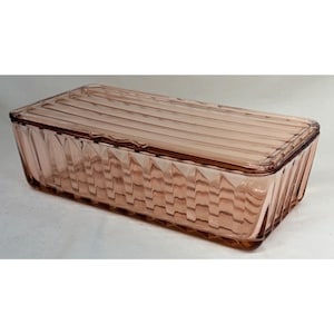 May include: A rectangular, light pink glass storage container with a matching lid. The exterior features a vertical ribbed pattern on the sides and a series of parallel horizontal lines across the top surface of the lid, creating a textured finish.