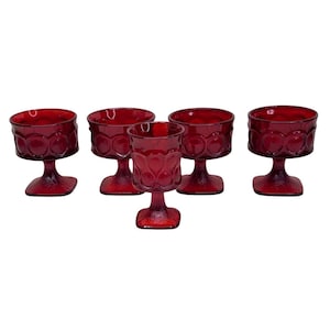 May include: A set of five ruby red glass goblets, each featuring a textured circular pattern around the bowl and a square base. These drinking vessels have a short, thick stem and a wide, rounded rim, showcasing a consistent deep red color.