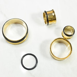 May include: A collection of gold-colored metal ear tunnels and plugs in various sizes, each featuring a smooth, polished finish. Three of the cylindrical pieces are fitted with thin black rubber O-rings, and one separate black rubber O-ring is included.