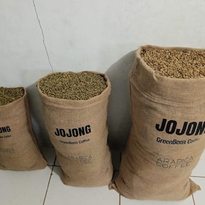 Könnte beinhalten: Drei beige Jutesäcke gefüllt mit rohen grünen Kaffeebohnen, angeordnet in aufsteigender Größe. Jeder Sack ist auf der Vorderseite mit dem schwarzen Aufdruck JOJONG GreenBean Coffee und ARABICA COFFEE versehen, die Bohnen sind oben sichtbar.