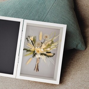 May include: A white shadow box frame containing a dried floral arrangement with cream, beige, and green tones. The bouquet features fluffy pampas grass, dandelion seed heads, fern leaves, and delicate skeletonised leaves, all tied with twine.