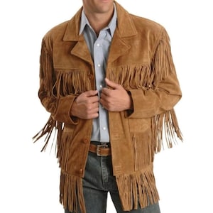 May include: A tan suede jacket featuring long fringe detailing across the chest, sleeves, and hem. The jacket has a collared neckline, a button-front closure, and is worn over a light blue button-down shirt and dark blue denim jeans.
