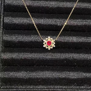 以下が含まれることがあります： 中央に丸い赤い宝石をあしらい、小さな透明な石で装飾された幾何学的な星型のフレームで囲まれたペンダントトップが付いたゴールドチェーンネックレス。ペンダントは繊細なゴールドのリンクチェーンから吊り下げられています。