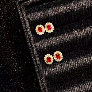 May include: Two pairs of stud earrings, each featuring a central oval-shaped red gemstone surrounded by a halo of small, clear, sparkling stones set in a metallic frame. The earrings are displayed in a black jewelry box with a satin lining.