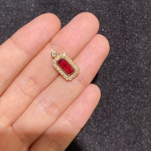 May include: A rectangular pendant featuring a deep red gemstone set in a gold-toned frame, surrounded by a border of small clear stones. A single small clear stone is attached to the top of the rectangular frame, connecting to a thin silver chain.