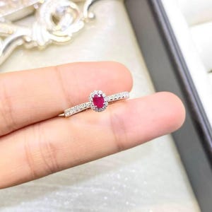 以下が含まれることがあります： 中央にオーバルカットの鮮やかな赤い宝石を配し、その周りを小さな透明な石のヘイローで囲んだシルバー調の指輪。リング部分は細身で、上半分に小さな透明な石が一列に埋め込まれています。