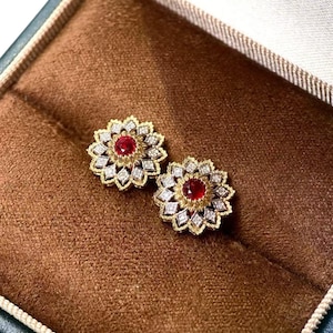 May include: A pair of floral-shaped stud earrings featuring a central round red gemstone surrounded by two concentric layers of small white stones set in yellow gold. The petals have a textured finish and the earrings are displayed in a brown box.