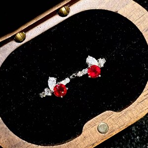 Puede incluir: Un par de pendientes plateados con una piedra preciosa redonda de color rojo vibrante como pieza central, acentuada por una piedra clara en forma de pera y piedras claras redondas más pequeñas, presentados en un joyero de madera oscura.