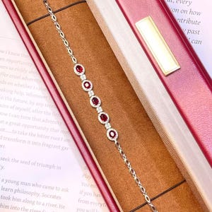 Peut inclure: Un bracelet argenté orné de cinq pierres rouges de taille ovale, chacune entourée d'un halo de petites pierres claires. La chaîne se compose d'anneaux ovales ouverts alternés et de petits motifs floraux, avec un fermoir métallique sécurisé.