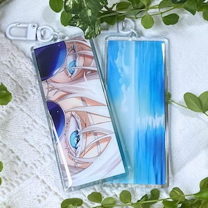 Könnte beinhalten: Zwei rechteckige Acryl-Schlüsselanhänger mit weißen Kunststoff-Karabinerverschlüssen. Ein Anhänger zeigt eine Anime-Illustration mit leuchtend blauen Augen hinter einer dunklen Sonnenbrille und weißem Haar. Der andere zeigt einen vertikalen blau-weißen Himmelsverlauf.