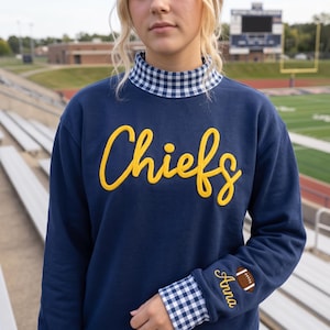 May include: A navy blue long-sleeve sweatshirt featuring a mock neck collar with a blue and white gingham pattern. The word Chiefs is embroidered in yellow script across the chest. The left sleeve displays the name Anna and a football graphic.