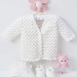 May include: A white crocheted long-sleeve cardigan with a button-front closure, a matching white crocheted bonnet, and a pair of white crocheted booties. A small pink plush cow rattle with a clear plastic ring containing colourful beads is included.