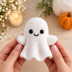 May include: A small, white, crocheted ghost figurine featuring a rounded head, two small arms, and a flared base. The ghost has two shiny, black, circular plastic eyes and a simple, thin, black embroidered smile centered on its face.