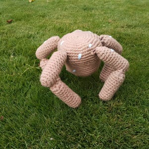 May include: A crocheted plush toy shaped like a spider with a round, light brown body and eight jointed legs. The surface of the body and legs features small, scattered white embroidered spots, giving the item a textured, handmade appearance.