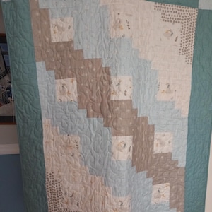 Puede incluir: Una colcha rectangular con un patrón diagonal de cuadrados en azul claro, beige y blanco. La tela muestra pequeñas ilustraciones de patos y follaje. La colcha está terminada con un borde sólido en color verde azulado y acolchado.