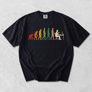 Pode incluir: Uma t-shirt preta de manga curta com uma impress&atilde;o gr&aacute;fica multicolorida que retrata a evolu&ccedil;&atilde;o humana, come&ccedil;ando por um primata e progredindo para uma pessoa sentada a uma secret&aacute;ria a trabalhar num computador com uma ch&aacute;vena.
