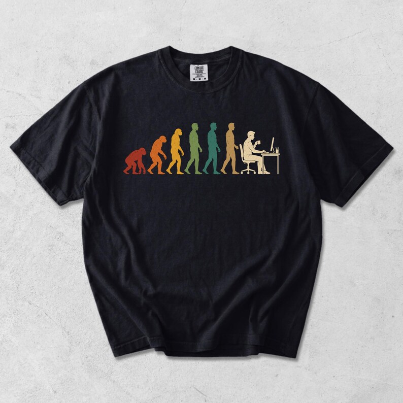 Pode incluir: Uma t-shirt preta de manga curta com uma impress&atilde;o gr&aacute;fica multicolorida que retrata a evolu&ccedil;&atilde;o humana, come&ccedil;ando por um primata e progredindo para uma pessoa sentada a uma secret&aacute;ria a trabalhar num computador com uma ch&aacute;vena.