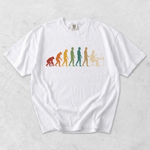 May include: A white short-sleeved t-shirt featuring a multi-colored graphic print on the chest. The design depicts the evolutionary progression from a primate to a human sitting at a desk working on a computer, rendered in shades of red, orange, yellow, green, blue, and tan.
