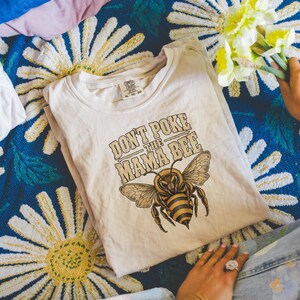 Può includere: Una t-shirt a maniche corte color crema con la grafica di un'ape a strisce marroni e gialle. Sopra l'ape, il testo "DON'T POKE THE MAMA BEE" è scritto in un carattere marrone stilizzato. La maglietta presenta un girocollo e un'etichetta.