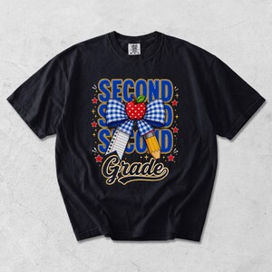 Può includere: Una t-shirt nera con un design grafico che riporta il testo SECOND GRADE. Il disegno include un fiocco a quadretti blu e bianco, una mela rossa con pois bianchi, un foglio di carta a righe, una matita gialla e piccole stelle rosse.