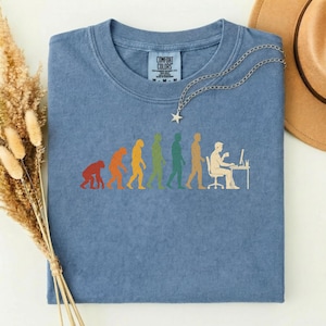 Pode incluir: Uma t-shirt azul de gola redonda com uma estampa gr&aacute;fica da evolu&ccedil;&atilde;o humana, progredindo de um primata para uma pessoa sentada a uma secret&aacute;ria com um computador. A t-shirt tem uma etiqueta Comfort Colors no pesco&ccedil;o e um colar com pendente de estrela prateado.