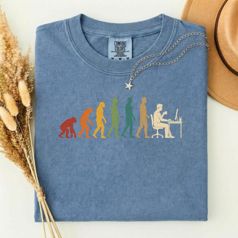Pode incluir: Uma t-shirt azul de gola redonda com uma estampa gr&aacute;fica da evolu&ccedil;&atilde;o humana, progredindo de um primata para uma pessoa sentada a uma secret&aacute;ria com um computador. A t-shirt tem uma etiqueta Comfort Colors no pesco&ccedil;o e um colar com pendente de estrela prateado.