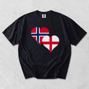 Può includere: Una t-shirt nera a maniche corte con due grafiche a forma di cuore sul petto. Il cuore più grande mostra la bandiera della Norvegia, mentre il cuore più piccolo sovrapposto mostra la bandiera dell'Inghilterra, entrambi con una finitura usurata.