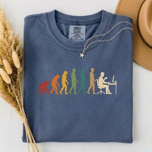 Pode incluir: Uma t-shirt azul de gola redonda com uma impress&atilde;o gr&aacute;fica multicolorida que mostra a evolu&ccedil;&atilde;o humana, come&ccedil;ando por um primata e terminando com uma pessoa sentada a uma secret&aacute;ria com um computador. A etiqueta diz COMFORT COLORS.