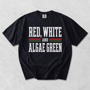 Könnte beinhalten: Ein schwarzes Kurzarm-T-Shirt mit dem weißen, abgenutzten Schriftzug "RED, WHITE AND ALGAE GREEN" mittig auf der Brust. Zwischen den Textzeilen befinden sich zwei horizontale Streifen, einer in Rot und einer in Grün, über und unter dem Wort "AND".