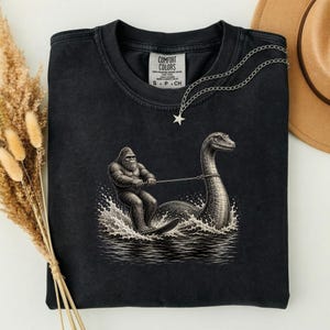 Maglietta con umorismo criptico "Bigfoot che fa sci nautico dietro a Nessie" PNG