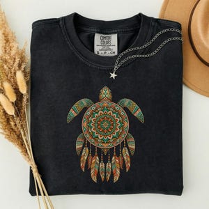 May include: A black crewneck t-shirt featuring a graphic of a sea turtle with a mandala-patterned shell and dangling feathers. The shirt has a Comfort Colors label at the inner neckline and is displayed with a silver star-shaped necklace.