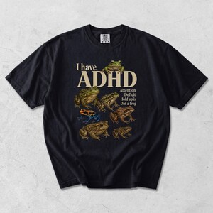 May include: A black short-sleeve t-shirt featuring a graphic of various frogs and the text I have ADHD Attention Deficit Hold up is Dat a frog. The design includes multiple frog species in shades of green, brown, and a bright blue and orange frog.