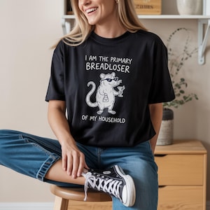 May include: A black short-sleeved t-shirt featuring a white graphic of an opossum wearing sunglasses, holding a small handbag and a martini glass. The text above and below the opossum reads, I AM THE PRIMARY BREADLOSER OF MY HOUSEHOLD.